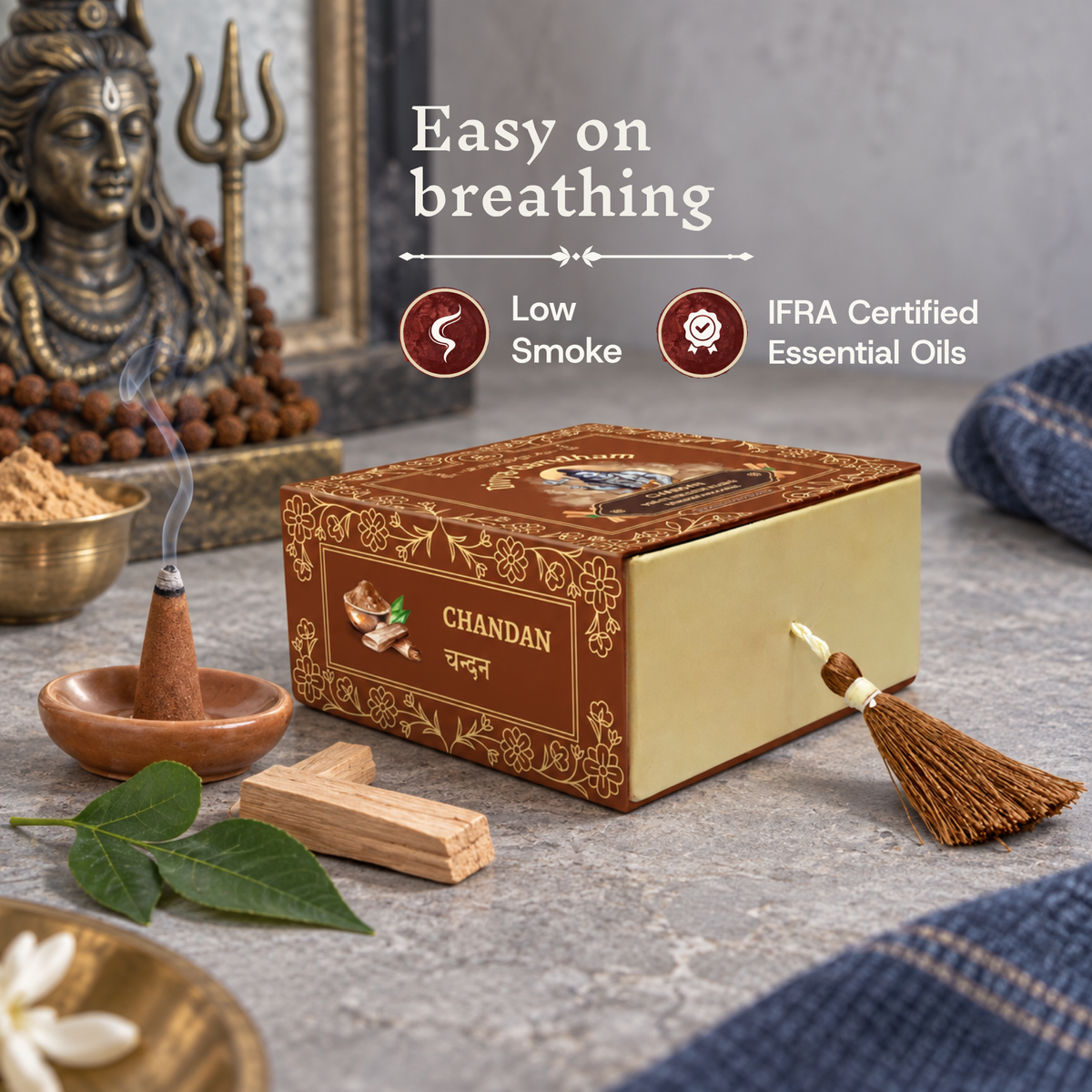 Lit Divyagandham Chandan incense cone highlighting easy breathing, IFRA certified essential oils, and low smoke, resting next to the product box, sandalwood pieces, and a Shiva statue.