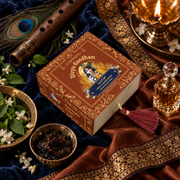 Overhead view of the Divyagandham Kasturi bambooless incense cones box featuring Lord Krishna, surrounded by a flute, peacock feather, and a lit brass diya on a blue velvet cloth.