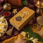 Overhead view of the Divyagandham Nagachampa bambooless incense sticks box featuring Maa Parvati, surrounded by white plumeria flowers, a lit diya, and kumkum on a dark red cloth.