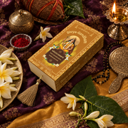 Overhead view of the Divyagandham Nagachampa bambooless incense sticks box featuring Maa Parvati, surrounded by white plumeria flowers, a lit diya, and kumkum on a dark red cloth.