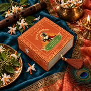 Overhead view of the Divyagandham Parijat bambooless incense cones box featuring Lord Krishna, surrounded by parijat flowers, a wooden flute, a peacock feather, and a lit brass diya on a blue cloth.