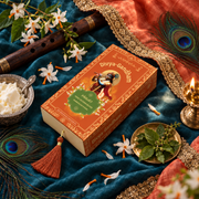 Overhead view of the Divyagandham Parijat bambooless incense sticks box featuring Lord Krishna, surrounded by parijat flowers, a wooden flute, peacock feathers, and fresh butter on a blue cloth.
